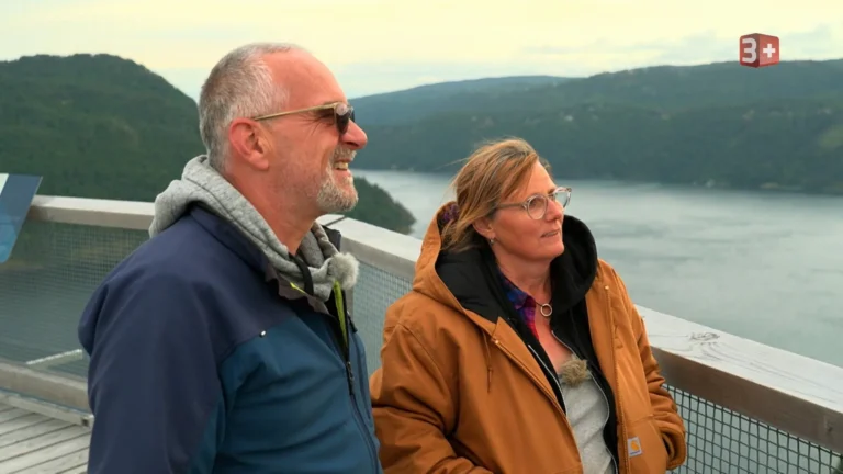 Two adults stand side by side on a wooden overlook railing, smiling at a lake and forested hills beyond.