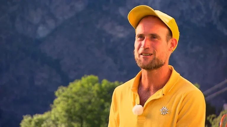 Man in a yellow polo and cap stands outdoors, mountains in the background, wearing a small clip-on microphone.