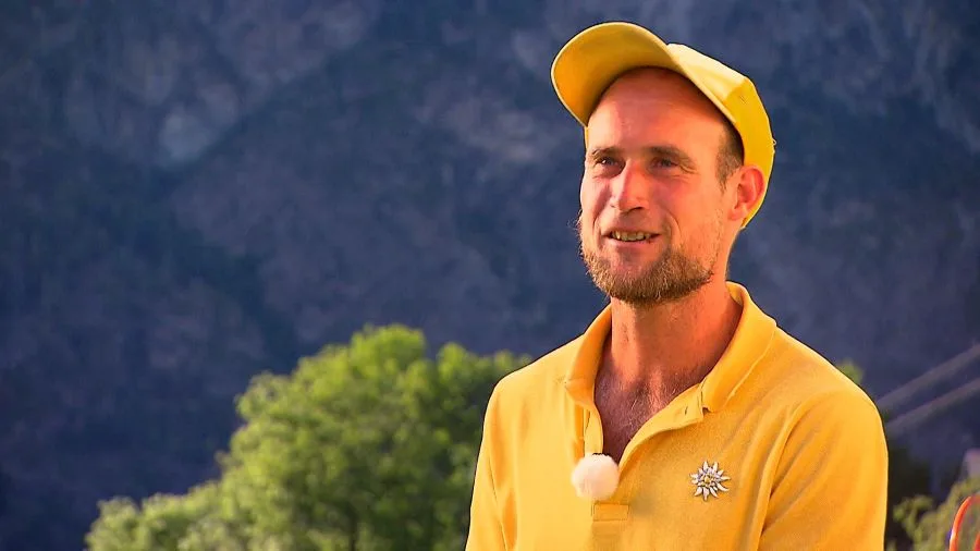 Man in a yellow polo and cap stands outdoors, mountains in the background, wearing a small clip-on microphone.