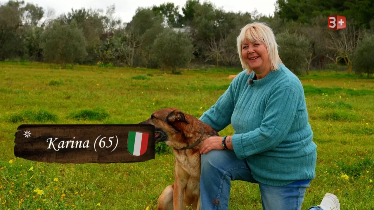 Older woman in a teal sweater kneels beside a brown dog in a grassy field, a wooden sign reads 'Karina (65)' with an Italian flag badge.