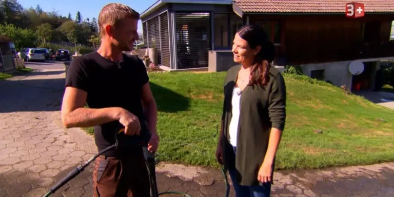 Two people stand on a driveway outside a house, chatting while the man holds a garden hose nozzle and a green hose.