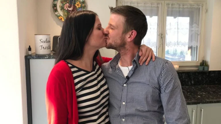 Couple sharing a kiss in a bright kitchen, arms around each other, woman wearing a striped shirt and red cardigan left of the man.