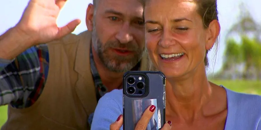 Smiling woman holding a smartphone for a selfie as a man waves behind her outdoors.