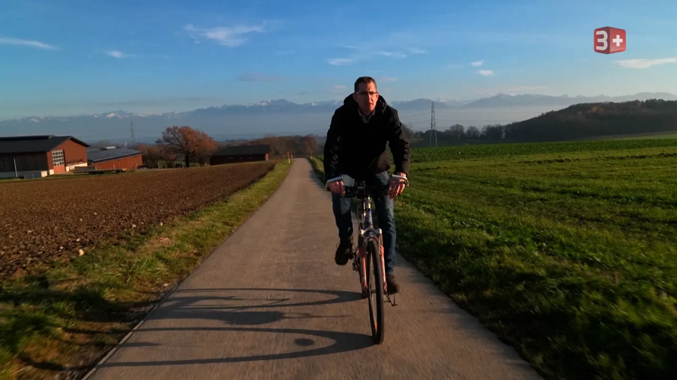 Person bicycling toward the camera on a rural paved path with fields and hills in the background.