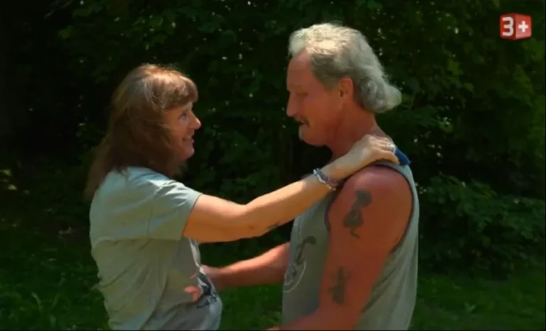 Two adults outdoors share a warm embrace, facing each other with smiles, against a green leafy background.