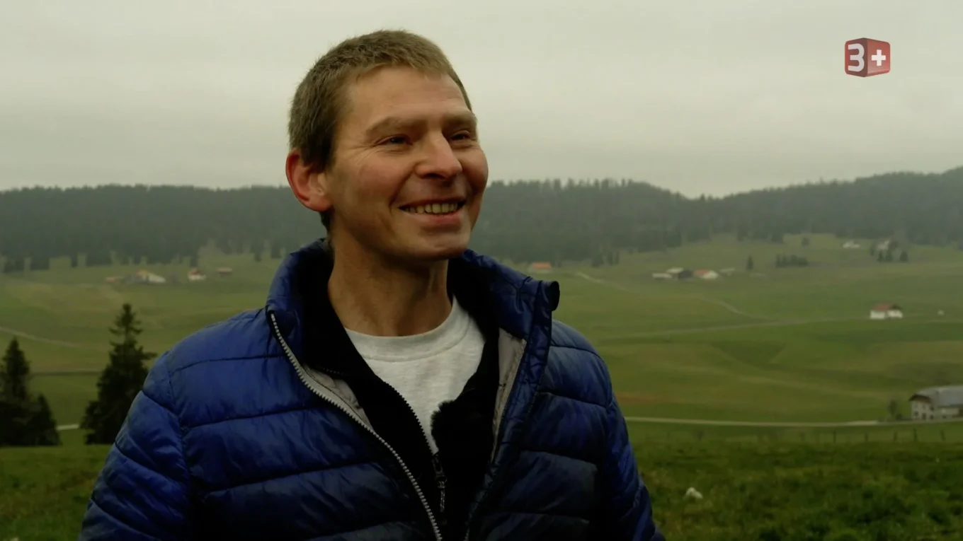 A smiling man wearing a blue puffer jacket stands outdoors in a grassy countryside with rolling hills and scattered houses in the distance.