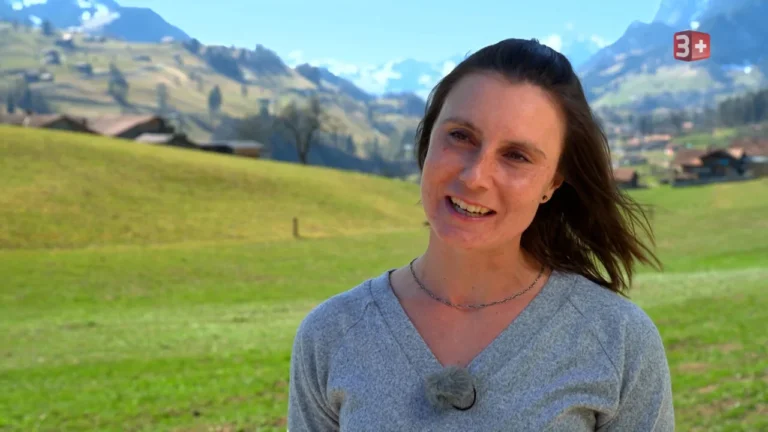 Smiling woman outdoors in a grassy field with mountains in the background; microphone clipped to her sweater.