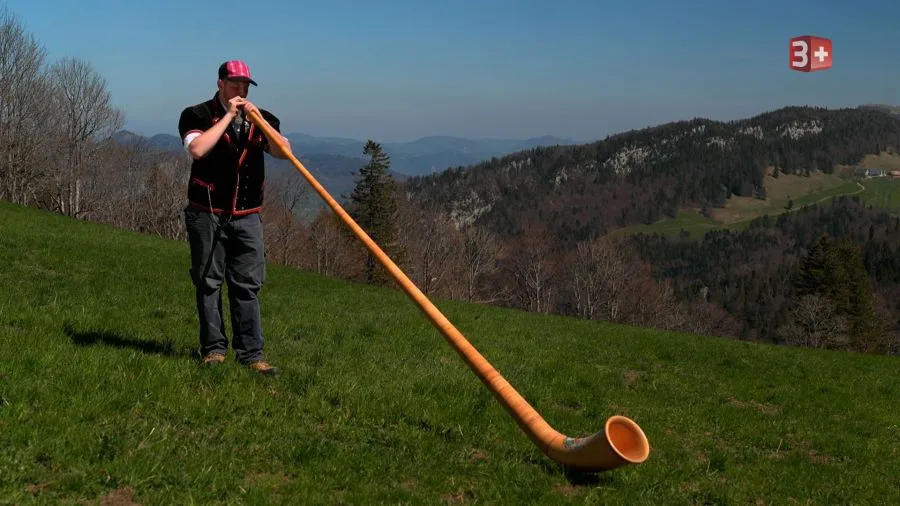Man wearing a pink cap stands on a grassy hillside and plays a long wooden horn toward the valley.