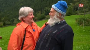 Two smiling older adults stand close together in a green hillside meadow; the woman wears an orange jacket and the man a dark striped sweater with a blue knit hat.