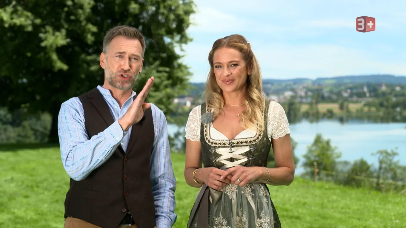 Man and woman in traditional Bavarian-style clothing stand outdoors near a lake, speaking to the camera with the scenery behind them.