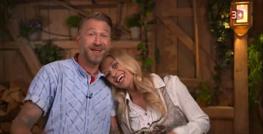 Smiling man and woman leaning together on a rustic wooden set with plants.