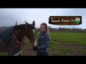 Woman in a blue quilted jacket stands beside a saddled horse in a green field; a wooden sign reads 'Marie Anna (40)'.